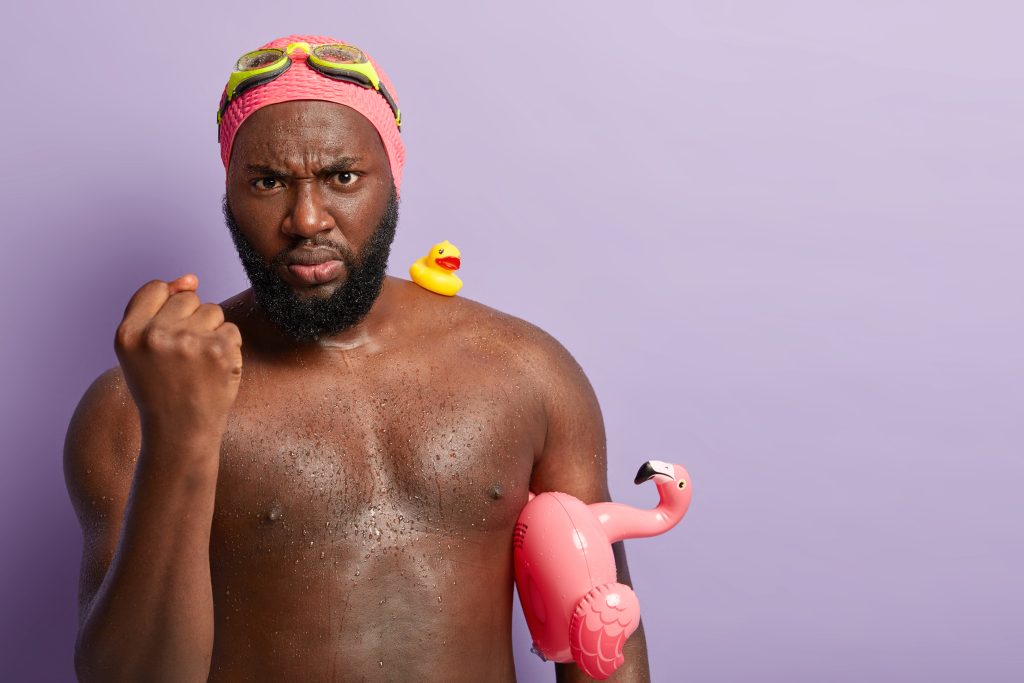 Man in swim cap and goggles holding inflatable flamingo, looking serious – meme marketing visual with playful tone.