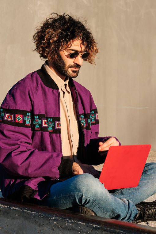 Man wearing a vintage purple patterned jacket and sunglasses, working on a bright red laptop outdoors. Represents retro design trends in modern branding, featured by The Smart Group.