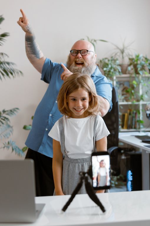 Social Listening & Micro-Virality, A cheerful young girl recording a video on a smartphone tripod while an excited adult stands behind her pointing and smiling, illustrating the playful, spontaneous content that often drives micro virality.