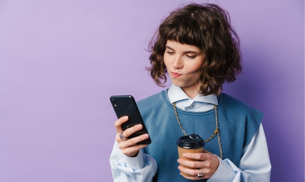 Social Listening & Micro-Virality. A young woman holding a coffee cup and looking thoughtfully at her phone against a purple background, representing social listening and micro virality trends.