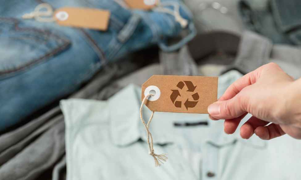 Greenwashing in the fashion industry, showing a hand holding a recycled cardboard clothing tag with a recycling symbol over folded garments, highlighting sustainability claims and environmental marketing.